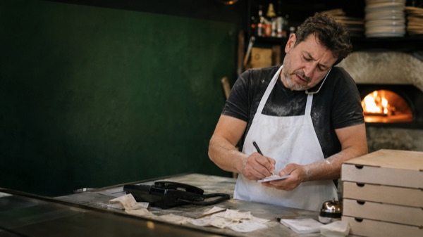 A pizza maker taking an order on the phone and writing it on a slip of paper, behind the counter of a pizzeria with the oven on and stacked boxes